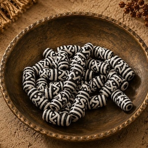 May include: A collection of cylindrical beads featuring a black and white geometric pattern, contained within a round, textured bronze-colored bowl. Each bead displays a repeating linear design with intricate markings across its surface.