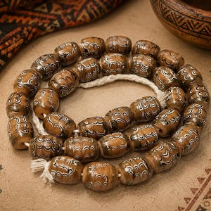 Könnte beinhalten: Eine Kette aus zylindrischen Glasperlen mit einem Marmormuster in Braun-, Schwarz- und Weißtönen. Die Perlen sind auf einer dicken weißen Schnur aufgereiht, kreisförmig angeordnet und weisen eine glänzende Oberfläche mit wellenförmigen Mustern auf.