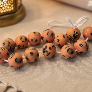 Puede incluir: Un collar de doce cuentas esféricas de color melocotón ensartadas en un cordón blanco. Cada cuenta presenta un patrón único de pequeñas marcas abstractas de color marrón oscuro y negro que parecen ojos estilizados sobre la superficie lisa.