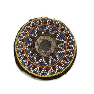 May include: A round, beaded, decorative piece with a central mirror. The beads are arranged in concentric circles of red, blue, yellow, and white.