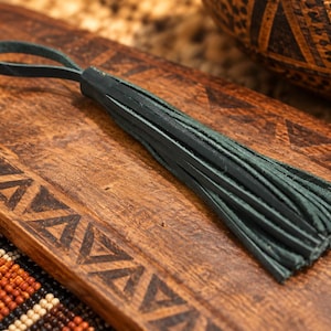 May include: A dark green leather tassel featuring a series of thin, vertical strips gathered at the top into a small, circular loop. The tassel is constructed from smooth, matte-finished leather material and displays a uniform deep forest green colour.