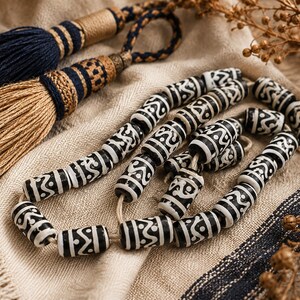 Puede incluir: Un collar de cuentas cilíndricas con un patrón geométrico en blanco y negro, acompañado de dos borlas decorativas en azul marino y beige con detalles de hilo dorado. Las cuentas están dispuestas en un bucle suelto sobre una tela texturizada.