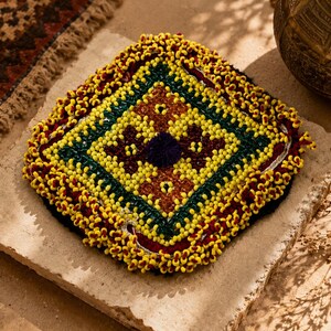 May include: A square textile coaster or mat featuring a central dark purple circular accent surrounded by concentric diamond patterns in yellow, dark red, and green. The entire perimeter is heavily embellished with dense yellow beadwork.