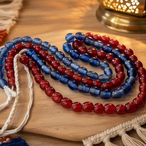May include: A prayer bead strand featuring alternating sections of translucent red and blue oval-shaped beads. The beads are strung on a white braided cord, with the ends of the cord left loose and visible at the side of the circular arrangement.