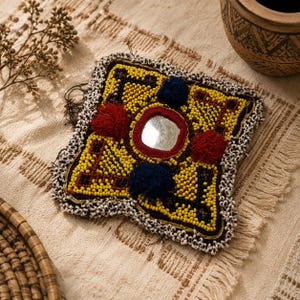Puede incluir: Un parche textil cuadrado con intrincados abalorios amarillos y negros, un espejo circular central enmarcado en rojo y cuatro pompones decorativos en rojo y azul oscuro. Los bordes están rematados con una cenefa de pequeñas cuentas blancas.