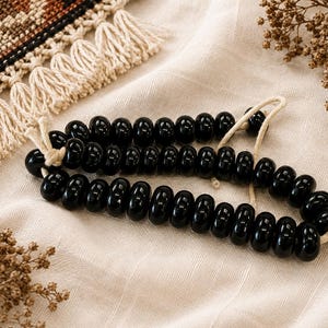 Puede incluir: Un collar de cuentas negras lisas y brillantes ensartadas en un cordón de color claro. Las cuentas son redondas y de forma uniforme, dispuestas en un bucle continuo. La superficie oscura y reflectante contrasta con el cordón de tono neutro.
