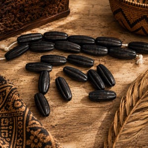 May include: A collection of elongated, oval-shaped black beads with vertical ridges. Some beads are strung together on a light-colored cord, while others are scattered loosely. The beads have a dark, matte finish and a uniform, symmetrical design.