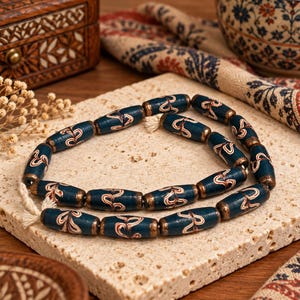 May include: A strand of cylindrical beads in a deep blue hue, featuring intricate white and red swirling patterns. Each bead is capped with a metallic bronze finish at both ends, and they are strung together on a light-coloured cord.