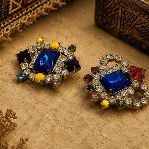 Op de afbeelding: Twee decoratieve broches met grote rechthoekige blauwe middenstenen omringd door kleinere heldere strass-steentjes en geaccentueerd met kleurrijke gele, rode, paarse en roze cabochons in een metalen frame met een textuur.
