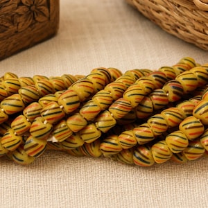 May include: A collection of small, barrel-shaped beads in a mustard yellow hue, each featuring diagonal stripes in black and burnt orange. The beads are strung together in multiple rows, creating a dense, textured cluster of uniform, cylindrical pieces.
