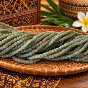 May include: A collection of numerous strands of small, matte, pale sage green beads arranged in a dense, horizontal pile. Each bead features a slightly irregular, rounded square shape with a smooth, muted finish, creating a uniform texture.