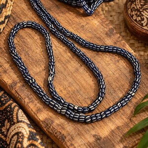 May include: A long strand of small, cylindrical beads featuring a dark navy blue base with thin, vertical white stripes. The beads are strung together in a continuous loop, displaying a uniform pattern and texture throughout the entire length of the necklace.