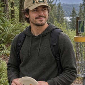 Puede incluir: Una gorra de béisbol de color tostado con un bordado de un jackalope lanzando un disco rojo frente a una cordillera. La gorra tiene una visera curva y una corona estructurada. El disco que se sostiene muestra un gráfico de una montaña.