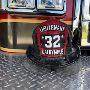May include: A black fire helmet featuring a red leather front piece with the text LIEUTENANT, the number 32, and the name DALRYMPLE in white lettering. The helmet has a curved brim and is secured with metal rivets on the front shield.