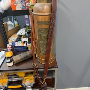 May include: A vintage copper fire extinguisher with a brass top and a large central plaque labeled QUEEN. A dark brown leather shoulder strap with the name BUFFKIN embossed in black letters is attached to the metal handle and side hardware.