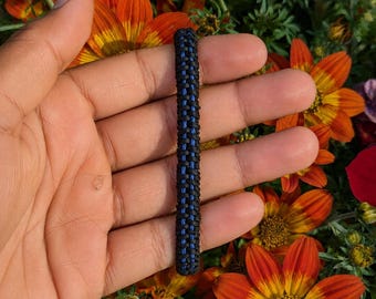 Op de afbeelding: Een paracord armband met een zwart vlechtpatroon en een dunne, verticale blauwe lijn in het midden. De armband is gemaakt van duurzaam koordmateriaal en toont een strak, ingewikkeld knoopontwerp over de gehele lengte van de band.