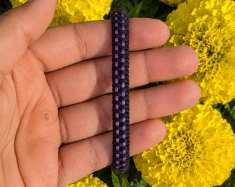Op de afbeelding: Een gevlochten armband met een centrale verticale streep van felpaars koord, geflankeerd door twee buitenste rijen van zwart koord, wat zorgt voor een getextureerd en consistent vlechtpatroon over de gehele lengte van het accessoire.