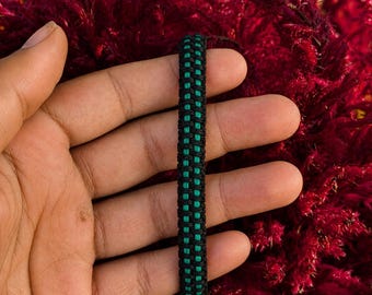 Op de afbeelding: Een gevlochten armband met een centrale verticale streep van felgroen koord, geflankeerd door twee parallelle rijen zwart koord. Het ontwerp bestaat uit een strak, herhalend knooppatroon dat een textuur en uniform uiterlijk geeft.