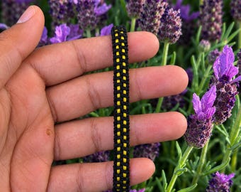 Op de afbeelding: Een gevlochten armband met een centrale verticale streep van felgeel koord, geflankeerd door twee buitenste rijen van zwart koord, wat een gestructureerd, rasterachtig patroon creëert over de lengte met een consistent knoopontwerp.
