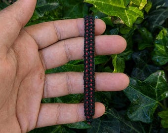 Op de afbeelding: Een gevlochten armband met een centrale verticale streep van felrood koord, geflankeerd door twee parallelle rijen zwart koord, gemaakt met een strak, ingewikkeld knooppatroon dat een gestructureerde, duurzame en smalle band vormt.