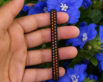 May include: A woven bracelet featuring a central vertical stripe of bright orange cord flanked by two parallel rows of black cord. The bracelet has a textured, braided appearance with a consistent pattern throughout its length.