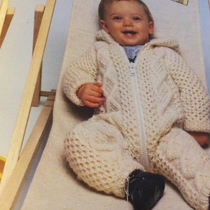 May include: A white knitted baby jumpsuit with a zipper closure and a cable knit pattern. The jumpsuit is worn by a baby sitting in a wooden chair.