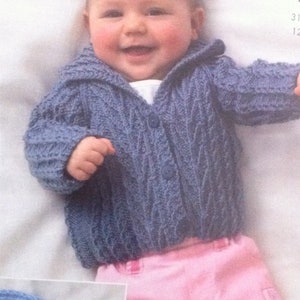 May include: A baby wearing a blue knitted cardigan with a cable knit pattern and a button closure. The cardigan has a collar and is worn over a pink shirt. The baby is smiling and has a light complexion.