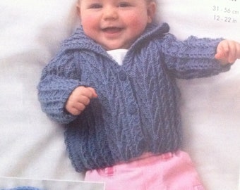 May include: A baby wearing a blue knitted cardigan with a cable knit pattern and a button fastening. The cardigan has a collar and is worn over a pink shirt. The baby is smiling and has a fair complexion.