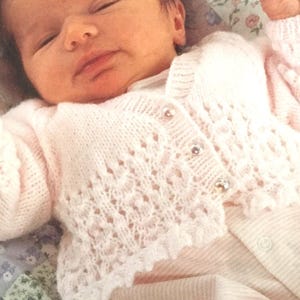 May include: A pink knitted baby cardigan with a delicate lace pattern and pearl buttons.