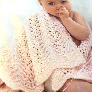 May include: A pink crocheted baby blanket with a delicate lace pattern. The blanket is draped over a baby, who is wearing a white hat.