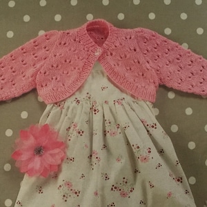 May include: A pink knitted baby bolero with a delicate openwork pattern. The bolero is worn over a white dress with a floral print.
