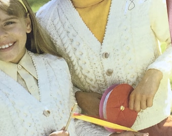 May include: Two children wearing white knitted cardigans with button closures. The cardigan on the left has a V-neck and the cardigan on the right has a round neck. The child on the right is holding a red and white spool of thread.