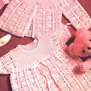 May include: A pink crocheted baby dress and matching cardigan with a delicate floral pattern. The dress has a tie at the waist and the cardigan has a button closure.