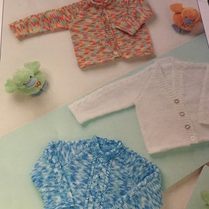 Puede incluir: Tres cárdigans de punto para bebé en diferentes colores y patrones. El primer cárdigan es un cárdigan multicolor con cierre de botones. El segundo cárdigan es blanco con cierre de botones. El tercer cárdigan es azul con cierre de botones.