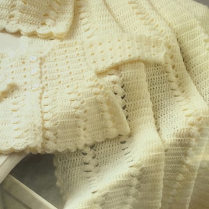 May include: A crocheted baby blanket and matching cardigan in a pale yellow color. The blanket is folded and the cardigan is laid on top. Both items are made with a delicate stitch pattern.