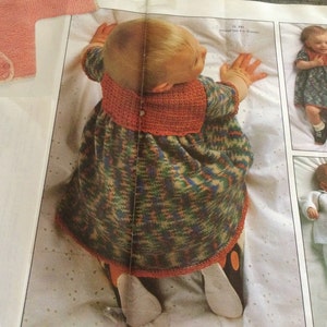 May include: A baby wearing a multi-colored knitted dress with a red and orange crocheted collar. The dress has a loose fit and is made with a textured yarn.