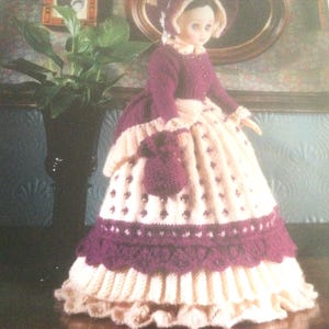 May include: A knitted doll wearing a white and purple dress with a matching hat and a small purple bag. The doll has a white face with painted features and brown hair.