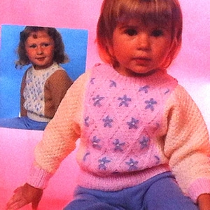 Könnte beinhalten: Ein junges Mädchen trägt einen rosa-weißen Strickpullover mit Blumenmuster. Der Pullover hat lange Ärmel und einen Rundhalsausschnitt. Das Mädchen lächelt und schaut in die Kamera.