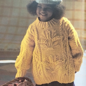 May include: A young girl wearing a yellow knitted sweater with a floral design. The sweater has a cable knit pattern and a round neckline.