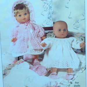 May include: Two dolls wearing knitted outfits. The doll on the left is wearing a pink crocheted coat and dress. The doll on the right is wearing a white crocheted dress. Both dolls are wearing pink shoes.