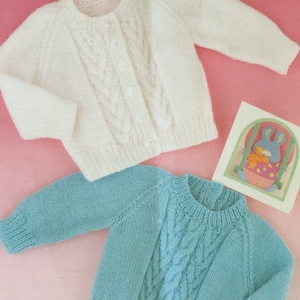 Könnte beinhalten: Zwei gestrickte Baby-Pullover, einer weiß und einer hellblau, mit einem Zopfmuster. Die Pullover liegen auf einer rosa Oberfläche.