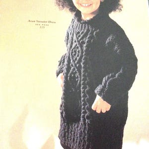 Peut inclure: Une robe pull en tricot Aran noire avec un motif en câble. La robe est portée par un jeune enfant. Le texte "Aran Sweater Dress See Page 115" est visible sur l'image.
