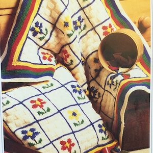 May include: A white crocheted blanket with a colorful floral pattern and a multi-colored border. The blanket is folded and draped over a wooden surface.