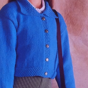 May include: A bright blue, knitted cardigan with a rounded collar and button closure. The cardigan has a textured pattern along the button line and hem. The buttons are a light brown color. The person is wearing a white shirt underneath and black and white striped pants.