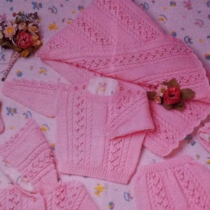 Puede incluir: Un conjunto de ropa de bebé de punto rosa que incluye un suéter, pantalones y una manta. El conjunto presenta un delicado patrón de punto de cable.
