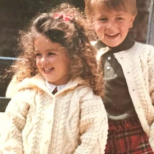 May include: Two children wearing white knitted cardigans. The girl has curly brown hair and is wearing a white collared shirt underneath her cardigan. The boy has short brown hair and is wearing a green shirt with a white design underneath his cardigan. He is also wearing a red and black plaid skirt.