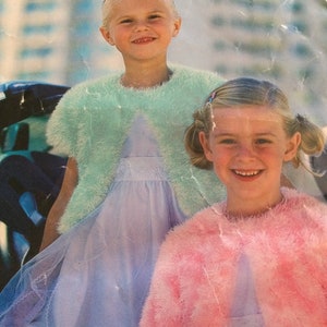 Op de afbeelding: Twee jonge meisjes dragen pluizige bolero's. Eén meisje draagt een lichtblauwe bolero en de ander een roze bolero. Beide meisjes lachen. Ze dragen jurken.