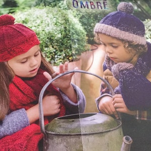 Puede incluir: Dos niños con gorros y bufandas de punto miran una regadera de metal. El gorro de la niña es rojo con un pompón blanco y su bufanda es roja. El gorro del niño es azul con un pompón gris y su bufanda es azul con una raya gris.