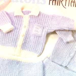 May include: A knitted baby outfit in a light blue color with white trim. The outfit includes a cardigan with a button closure and a matching pair of pants.