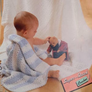 Peut inclure: Un bébé est enveloppé dans une couverture en crochet rayée blanche et bleue. Le bébé regarde un ours en peluche. La couverture est étiquetée "Super-Quick Crochet !"